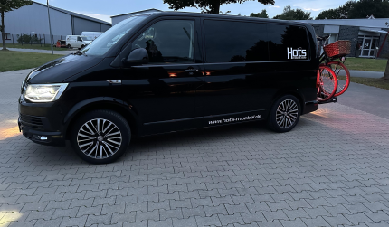 Schwarzer Campingbus mit Fahrradträger und Hots-Logo auf einem Firmenparkplatz