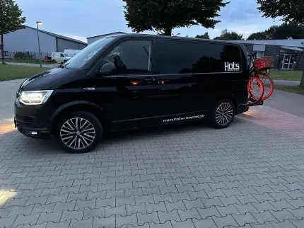 Schwarzer Campingbus mit Fahrradträger und Hots-Logo auf einem Firmenparkplatz