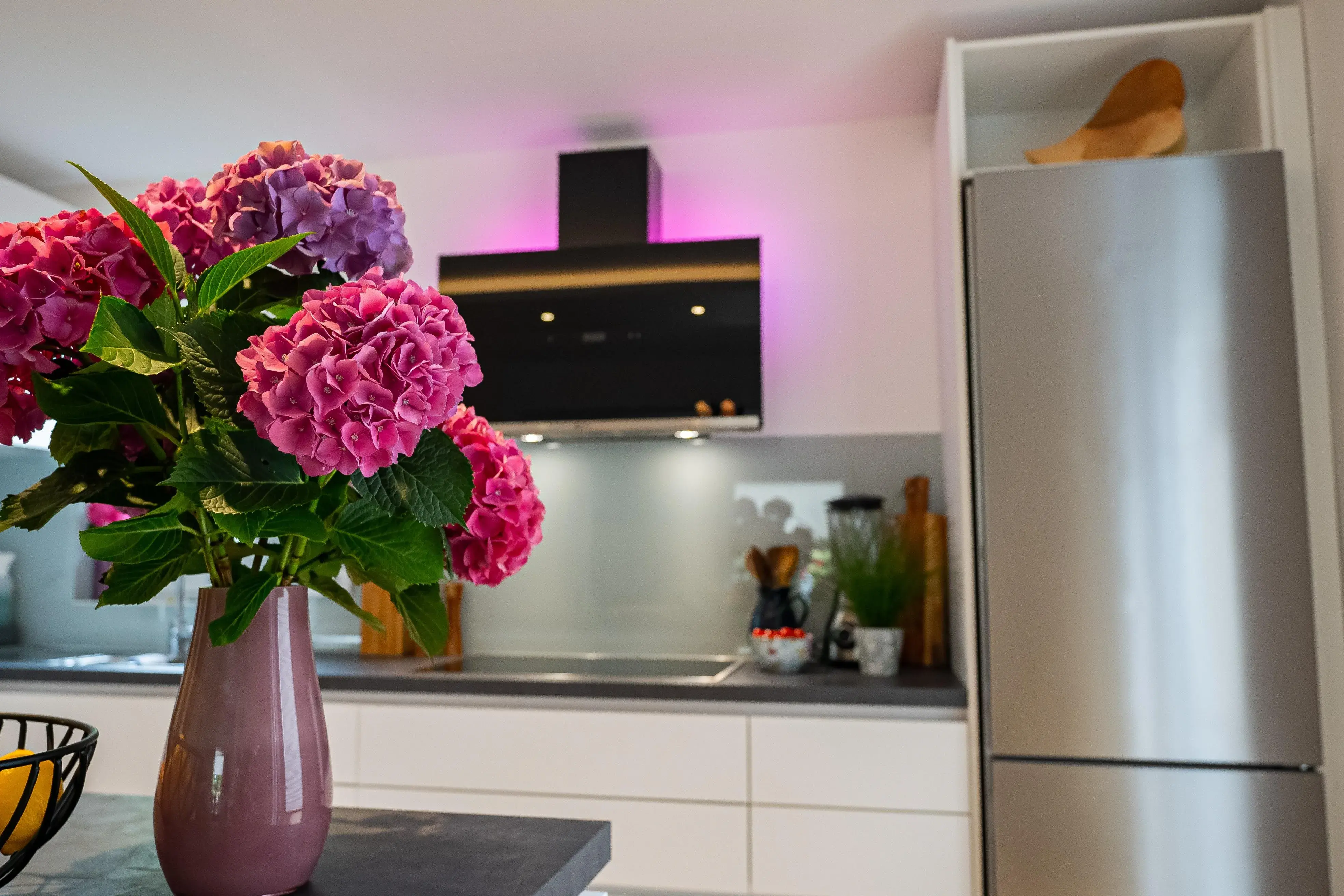 Moderne weiße Küche mit dunkler Arbeitsplatte, Edelstahl-Kühlschrank und rosa Hortensien