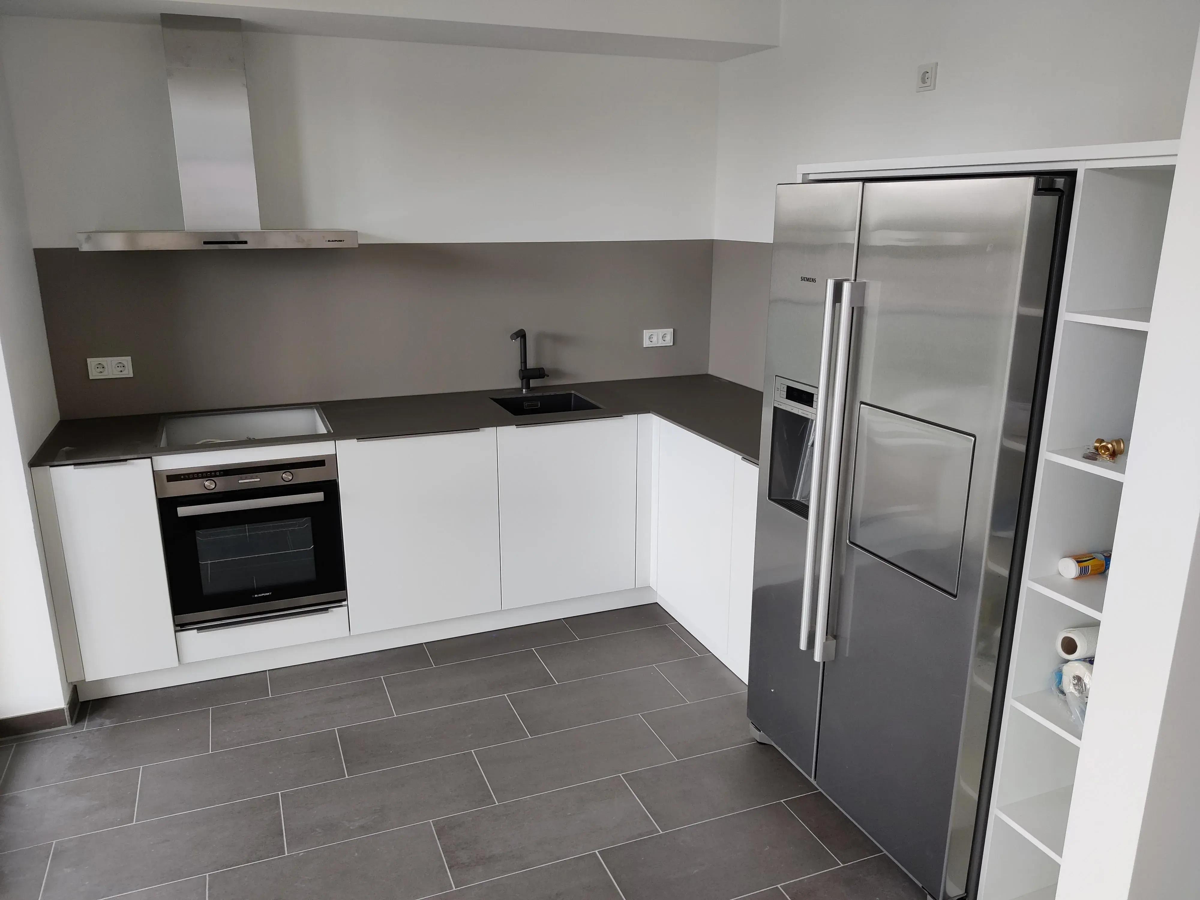Moderne weiße Winkelküche mit dunkler Arbeitsplatte, Einbaubackofen und Side-by-Side-Kühlschrank